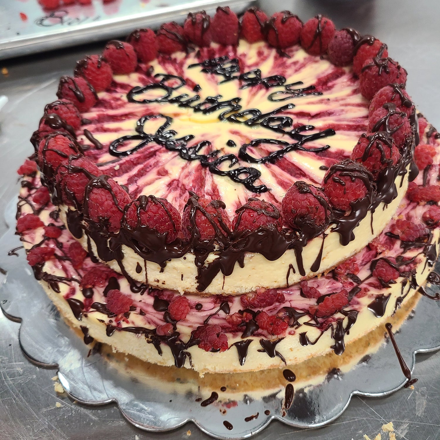 Elegant two-tier raspberry swirl cheesecake with fresh raspberries and chocolate drizzle, ideal for corporate events, celebrations, and special occasions.